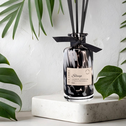 A black tortoiseshell diffuser bottle with black reeds on a white platform with green leaves in the background