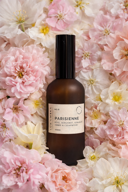 A room spray in an amber coloured bottle called Parisienne on a bed of pale pink flowers.