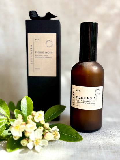 A brown frosted glass bottle of FIGUE NOIR room spray with a black label and cap, next to a black gift box wrapped with a black grosgrain ribbon, and a bunch of white flowers with green leaves.