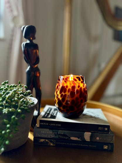 A lit candle in a tortoiseshell patterned glass jar on a stack of books, with a sculpture and a plant in the background.