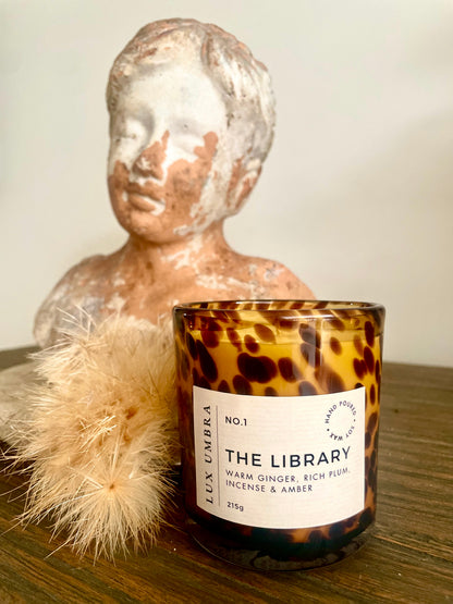A candle with a tortoiseshell glass jar, black lid, and label reading 'THE LIBRARY', alongside a decorative bust and a fluffy white seed pod