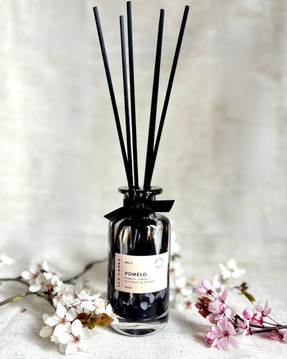 A black tortoiseshell diffuser called Pomelo, with black reeds and adorned with a black ribbon. De against a beige background and spring blossom.