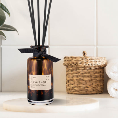 A brown tortoiseshell reed diffuser with a ripe black fig scent, placed on a white surface, with a wicker basket and white towels in the background.