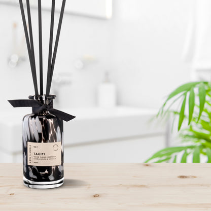 A black reed diffuser with a tortoiseshell patterned glass jar, six black fibre reeds, and a label reading 'TAHITI'. Presented on a wooden surface with a blurred background.