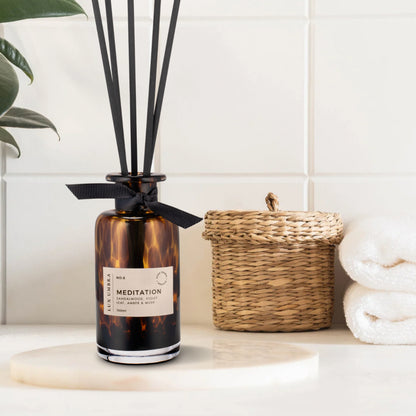 A brown tortoiseshell glass diffuser with black reeds and a label saying Meditation, next to rolled up towels 