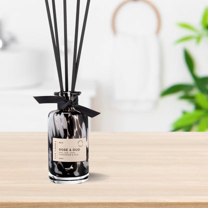 A black tortoiseshell glass jar with a label reading 'ROSE & OUD', decorated with a black ribbon and accompanied by six black fibre reeds. Sitting on a table in a bathroom.