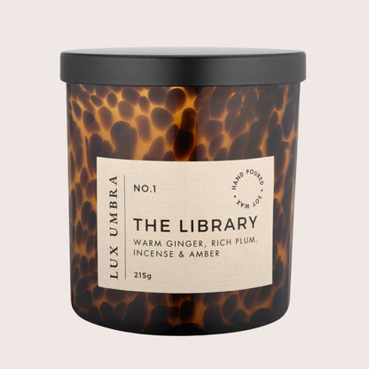 A soy and coconut wax candle in a tortoiseshell glass jar with a black lid, labelled 'THE LIBRARY' with warm ginger, rich plum, incense, and amber fragrance notes.