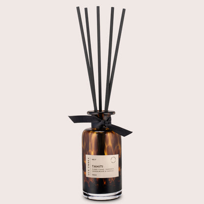 A brown reed diffuser with a tortoiseshell patterned glass jar, six black fibre reeds, and a label reading 'TAHITI'.On a beige  background.
