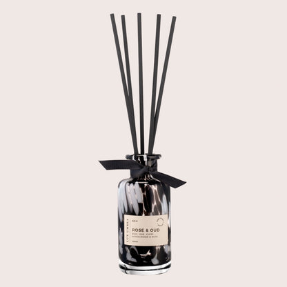 A black tortoiseshell glass jar with a label reading 'ROSE & OUD', decorated with a black ribbon and accompanied by six black fibre reeds.
