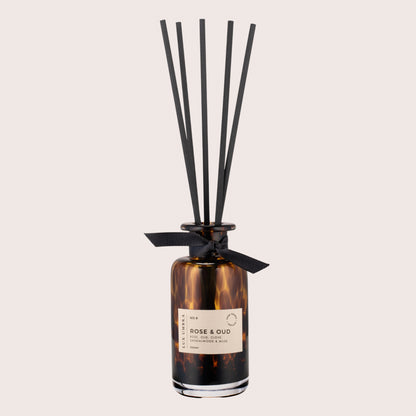 A brown tortoiseshell glass jar with a label reading 'ROSE & OUD', decorated with a black ribbon and accompanied by six black fibre reeds.