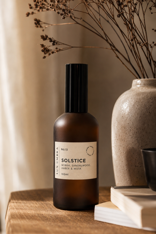 A brown amber glass bottle of Solstice room spray by Lux Umbra with a black cap and cream label. Styled on a wooden surface next to a ceramic vase with dried branches and books.
