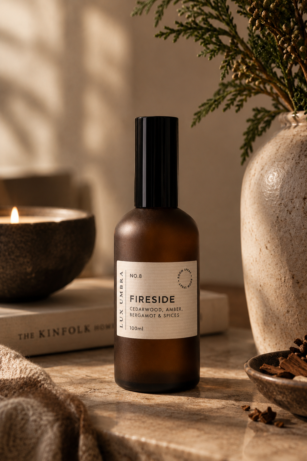 An amber glass room spray bottle with a black cap and a cream label reading 'FIRESIDE' and 'Cedarwood, Amber & Bergamot & Spices' by the brand Lux Umbra styled on a wooden surface with a lit candle, a book, and a textured ceramic vase in the background.