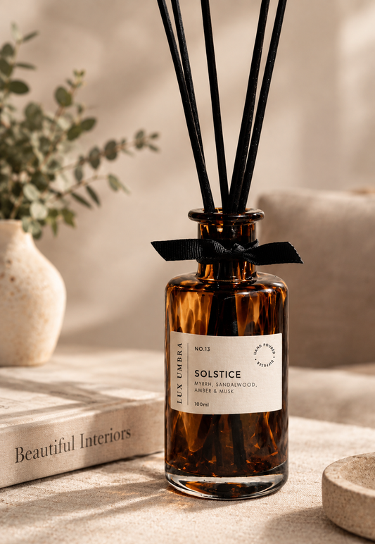 Brown tortoiseshell reed diffuser bottle with black reeds, a black ribbon bow, and a white label reading 'SOLSTICE' and 'LUX UMBRA', styled on a tabletop with a book and soft neutral background.
